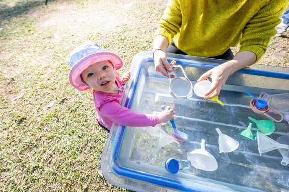 a toddler stands at a water table with a caregiver