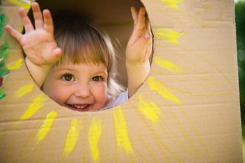 A young girl peeks her face out of a home made cardboard box, demonstrating sustainable, recycle play